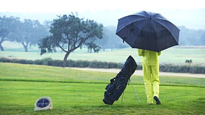 A golfer waits on a tee in full waterproofs and under an umbrella